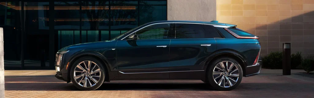 This image features a side view of the 2026 Cadillac Lyriq SUV, showcasing its sleek, modern design with large wheels, parked in front of a minimalist, reflective building.