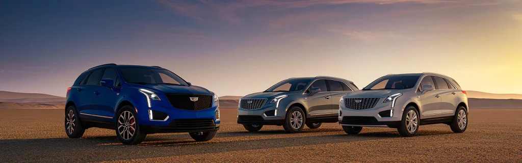 The image features three Cadillac XT5 SUVs displayed in a desert landscape during sunset. The vehicles are positioned from left to right: a blue, silver, and light gray model.