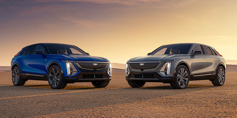 Two sleek, modern Cadillac LYRIQ—one blue, one gray—are parked side by side on a desert landscape at sunset, highlighting their stylish design and elegance.