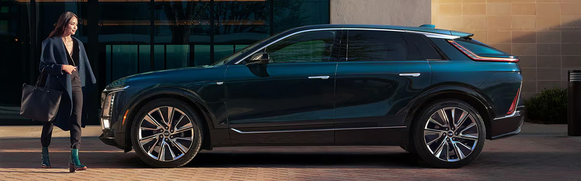 A woman walks towards a 2026 Cadillac LYRIQ parked in front of a modern building.