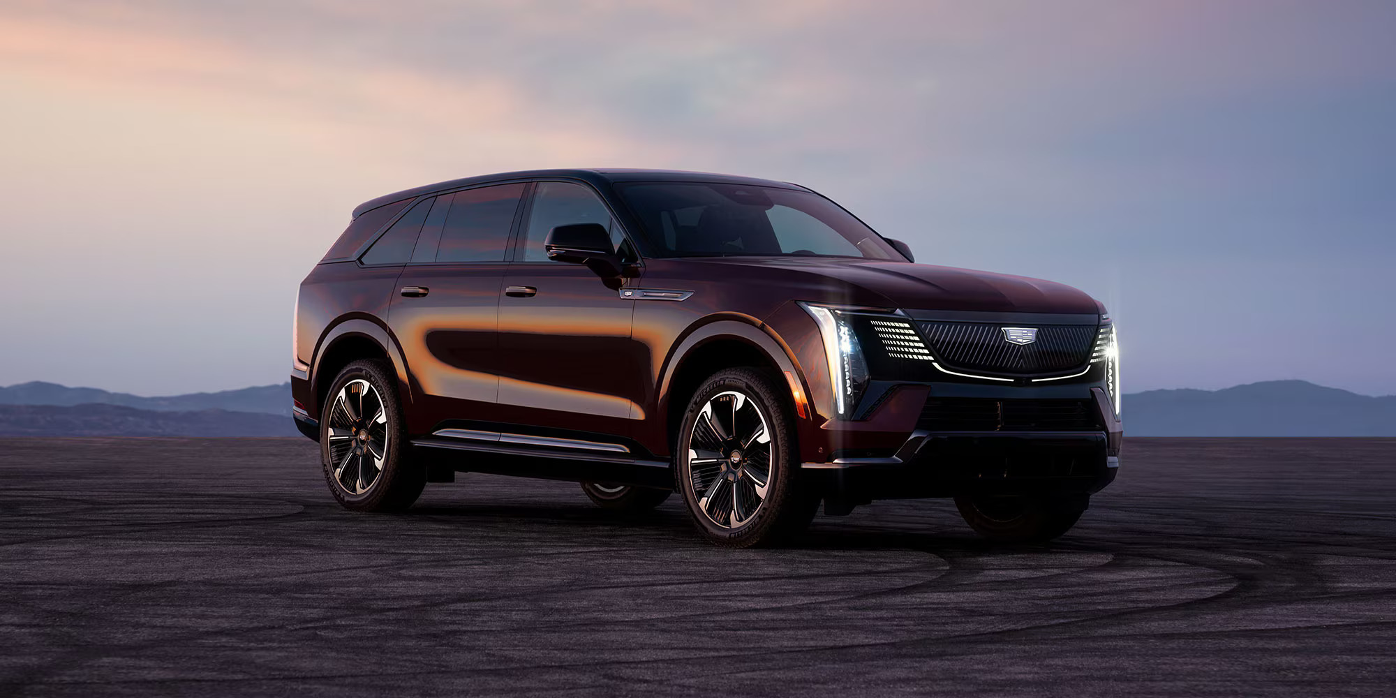 2026 Cadillac Escalade IQ luxury electric SUV in dark red with illuminated grille at sunset.